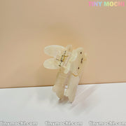 The Iridescent Bunny Hair Clip | Cute Hair Claw for Thick Hair by Tiny Mochi stands upright on a white surface, its translucent bunny face design glows subtly. Behind it, pastel TINY MOCHI text and tinymochi.com repeat on the beige background.