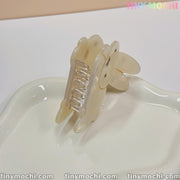 An Iridescent Bunny Hair Clip for thick hair from Tiny Mochi, with a strong grip, sits upright on a glossy white ceramic tray. The image shows tinymochi.com at the bottom and TINY MOCHI in rainbow colors at the top right.