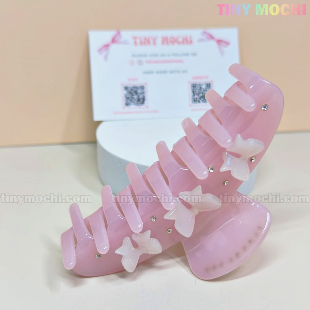 The Pink Bow Claw Clip by Tiny Mochi features 3D bows, small white butterfly accents, sparkling rhinestones, and is made from eco-friendly acetate. Displayed on a beige background with a white round base beside a Tiny Mochi card.