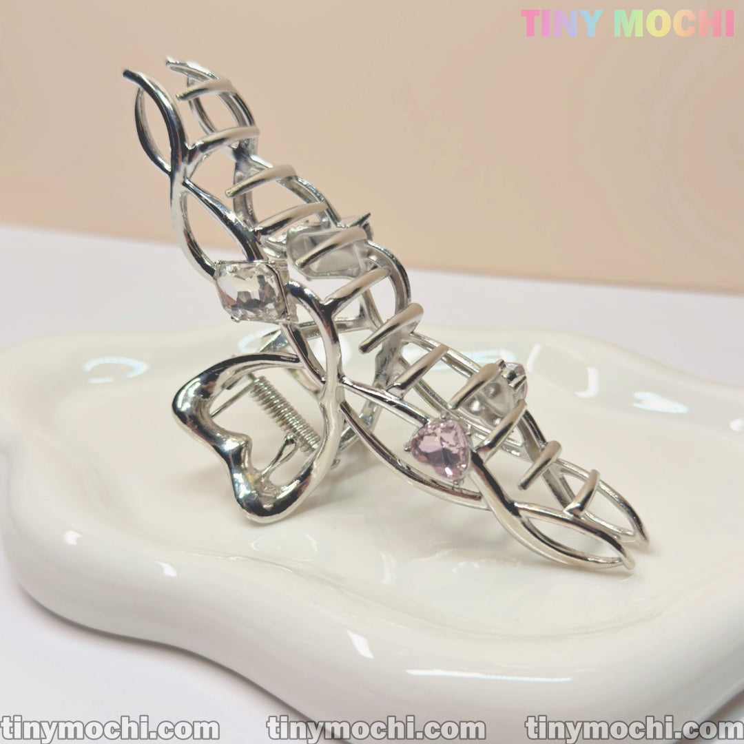 The Silver Crystal Heart Hair Claw by Tiny Mochi features a geometric, cage-like design with crystal heart accents. Shown on a glossy white dish, the pastel Tiny Mochi logo and website text are visible in the background.