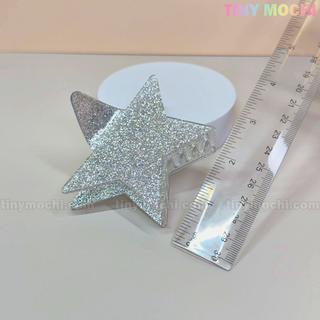 The Tiny Mochi Star Hair Claw Clip—Large Glitter Star Clip for Thick Hair—shaped like a silver star, sparkles beside a transparent ruler and white round object. This Y2K acetate hair accessory measures about 8 cm long.
