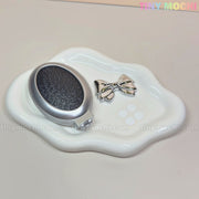 The Tiny Mochi Oval Silver Folding Travel Mirror Combâa compact pocket-sized brush with built-in mirrorârests on a wavy-edged white dish atop a beige surface, accompanied by a shiny silver bow hair clip.