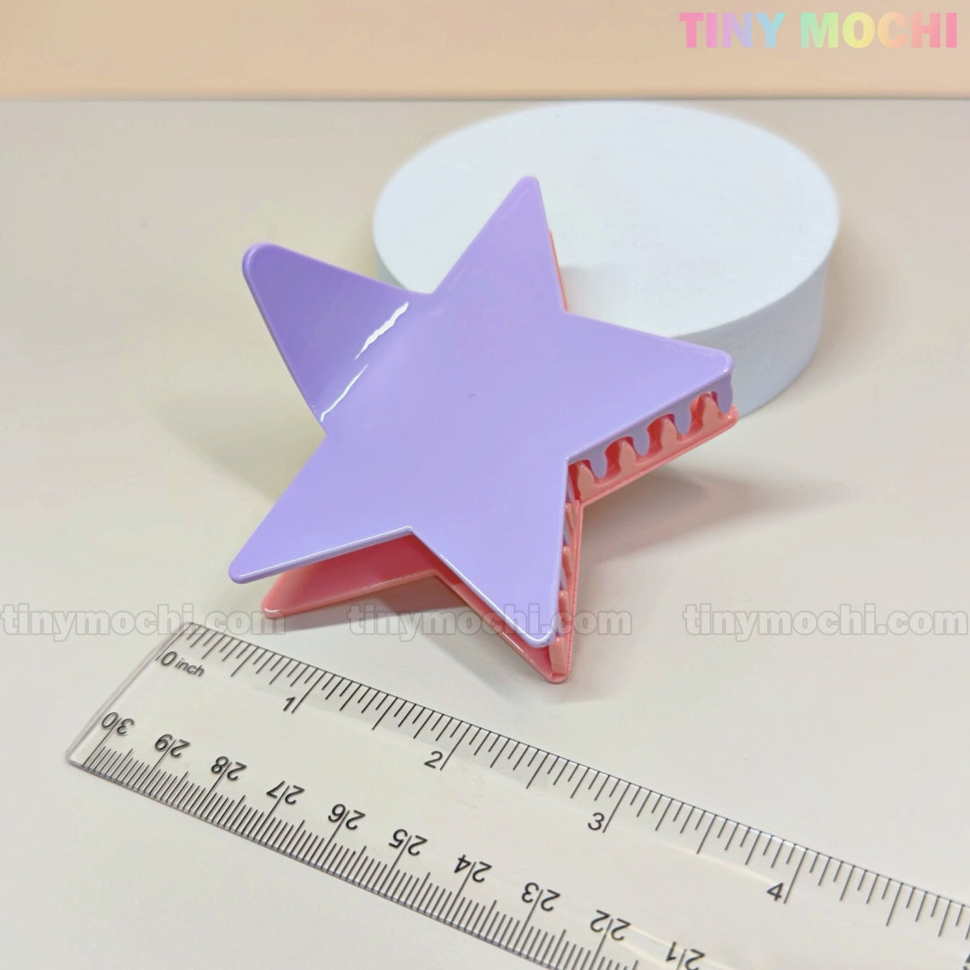 A pastel purple and pink Star Hair Claw Clip—Large Glitter Star Clip for Thick Hair by Tiny Mochi rests near a ruler and a white round object on a beige surface. TINY MOCHI appears in the top right corner.