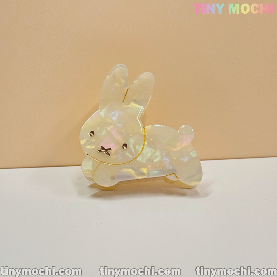 The Iridescent Bunny Hair Clip by Tiny Mochi, a shiny bunny-shaped hair claw with holographic glow and gold accents, rests on a white surface with a beige background. Tiny Mochi appears in pastel colors at the top right.