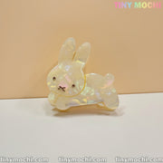 The Iridescent Bunny Hair Clip by Tiny Mochi, a shiny bunny-shaped hair claw with holographic glow and gold accents, rests on a white surface with a beige background. Tiny Mochi appears in pastel colors at the top right.