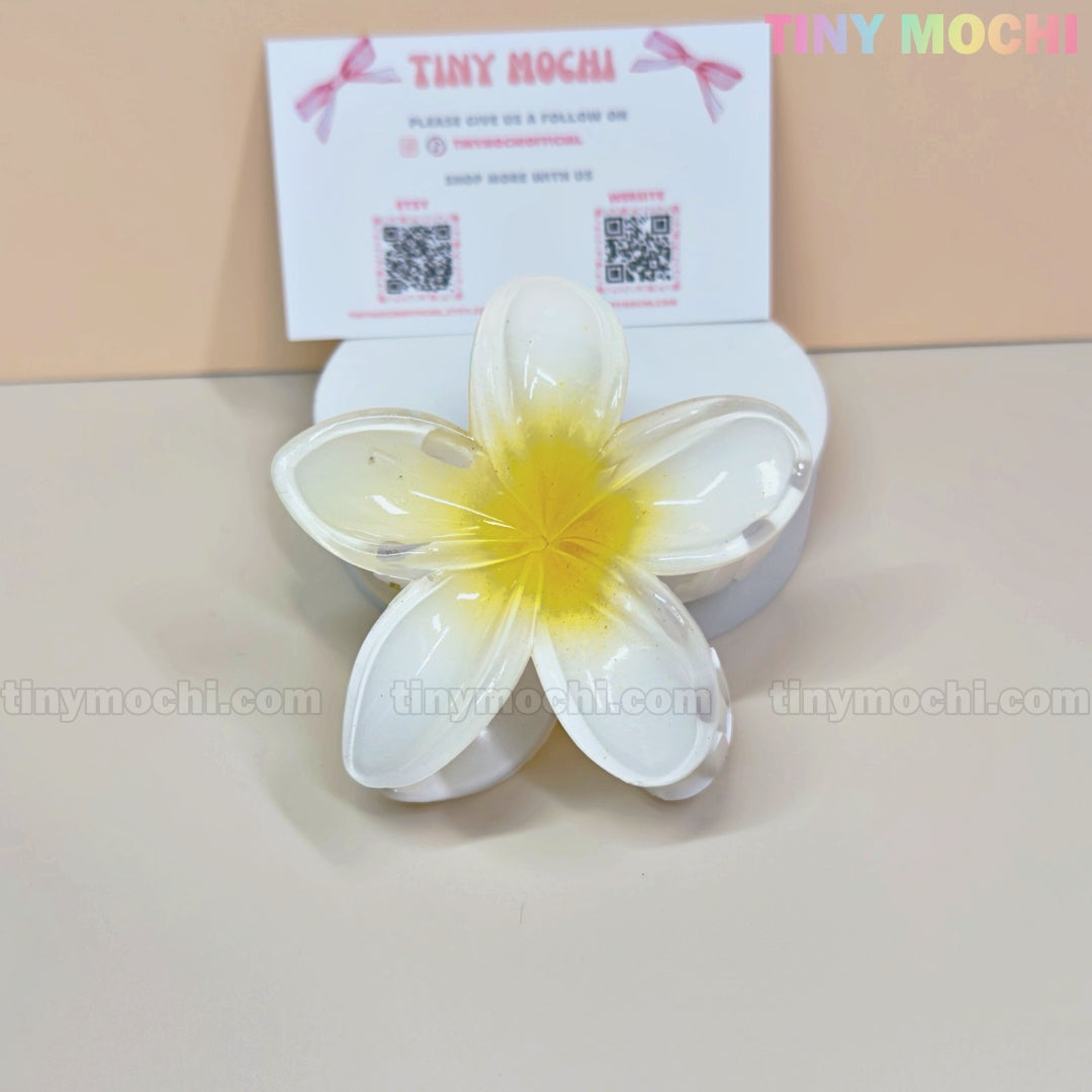 A colorful Plumeria Flower Hair Claw from Tiny Mochi rests on a white surface, with a branded card behind—perfect for fans of stylish summer hair accessories and floral clips for women and girls.
