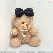 The Tiny Mochi Plush Teddy Bear Keychain with Bow and Pearl Necklace sits against a white background, featuring heart and star accents. Ideal as a cute bag charm, car key accessory, or stylish gift for girls. tinymochi.com appears below.