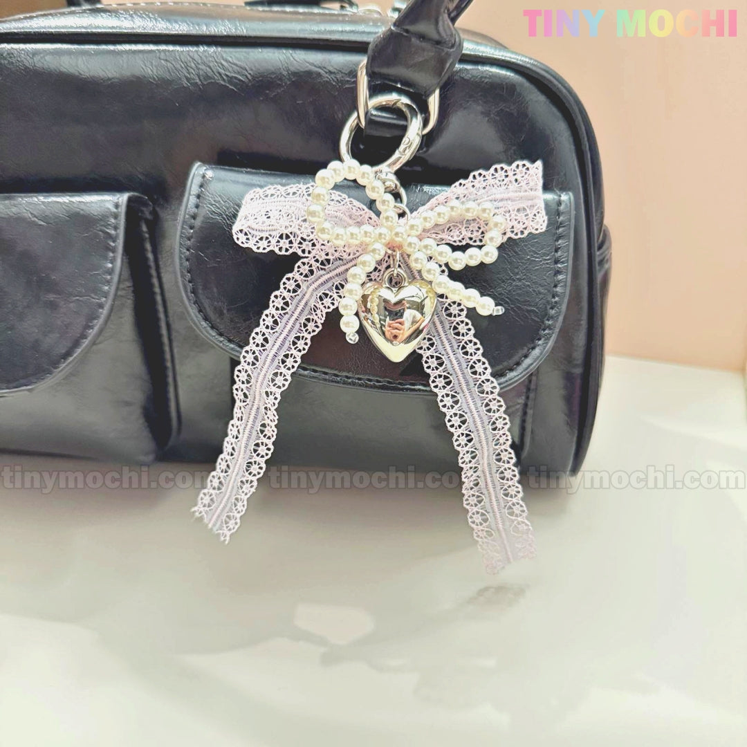 The Kawaii Lace Bow Keychain with pearl beads & a silver heart charm by Tiny Mochi decorates a black handbag, shown against a soft background with TINY MOCHI in the top right corner. Perfect as a bag charm, phone strap, or car mirror accessory.