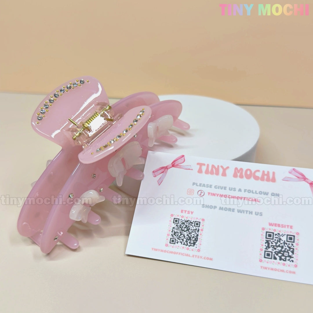 The Pink Bow Claw Clip by Tiny Mochi, made from eco-friendly acetate and decorated with rhinestones, rests on a round white platform. In front, a Tiny Mochi card with QR codes for Etsy and the website sits against pastel colors.