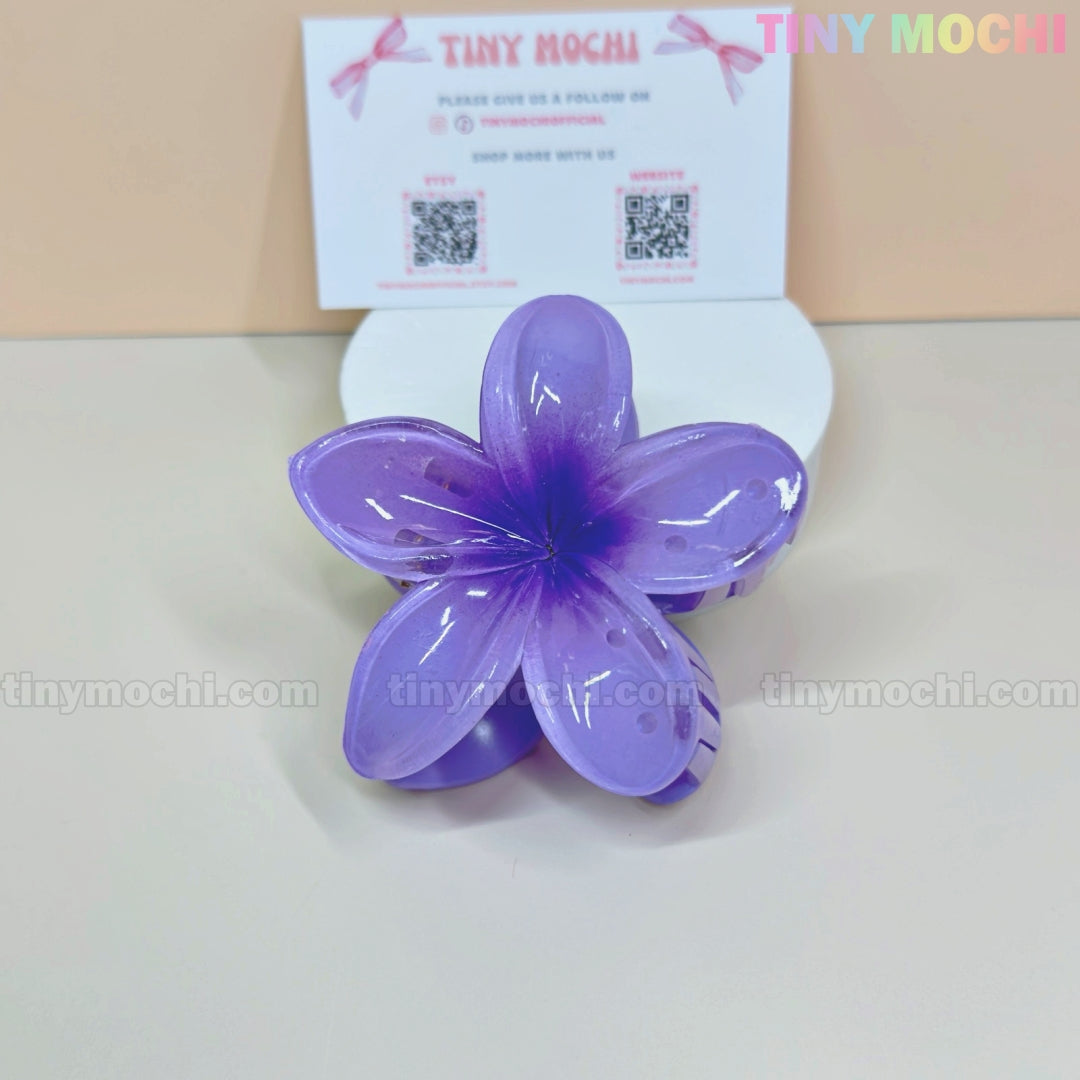 The Plumeria Flower Hair Claw from Tiny Mochi—a colorful floral clip for women & girls—is shown on a light surface with a Tiny Mochi business card and QR codes in back. This stylish summer accessory offers a strong grip and chic look. Background: pale beige.