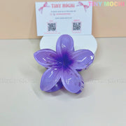 The Plumeria Flower Hair Claw from Tiny Mochi—a colorful floral clip for women & girls—is shown on a light surface with a Tiny Mochi business card and QR codes in back. This stylish summer accessory offers a strong grip and chic look. Background: pale beige.