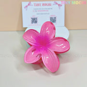 The Plumeria Flower Hair Claw by Tiny Mochi—a colorful floral hair clip for women and girls—rests on a white surface with a Tiny Mochi business card, making a stylish summer accessory for flower hair clip lovers.