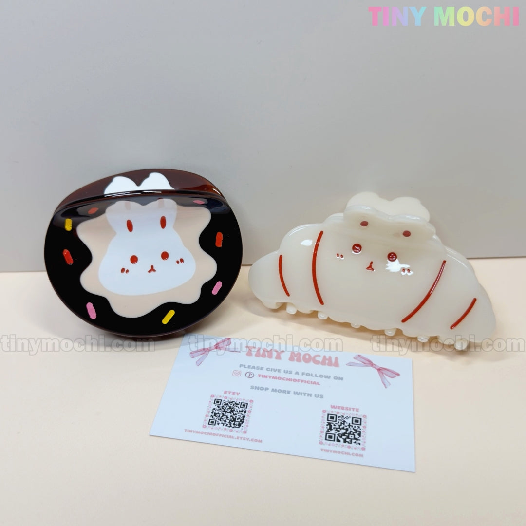 Two Kawaii Bunny Hair Claw Clips by Tiny Mochi—choose a Donut Sprinkle or Croissant design. Both cute acrylic accessories are shown on a light surface with a QR-coded card and pastel Tiny Mochi branding above. Perfect for any hairstyle!.