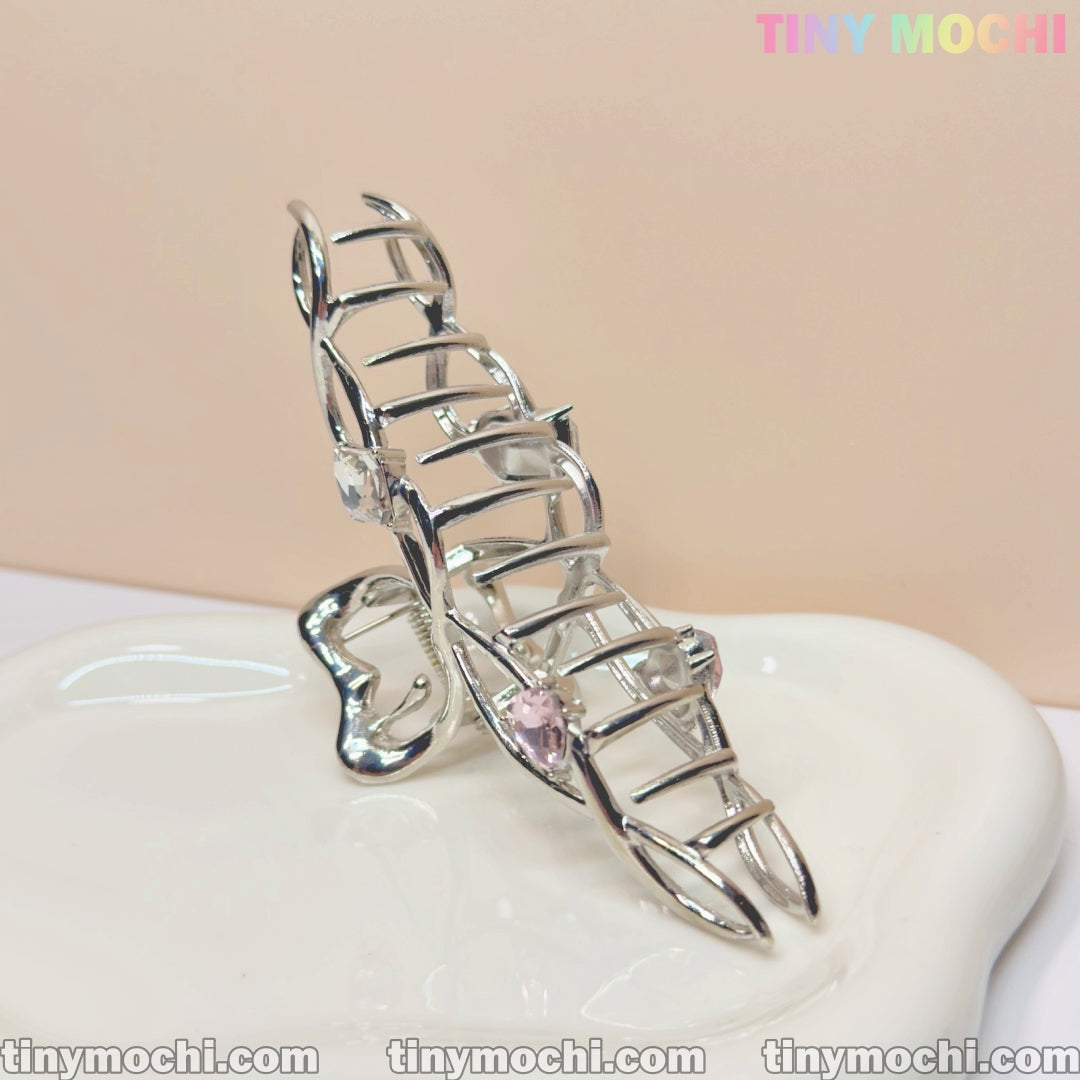 The Tiny Mochi Silver Crystal Heart Hair Claw, an elegant metal hair clip for women, is displayed upright on a white dish with a light beige background and the pastel Tiny Mochi logo in the top right corner.