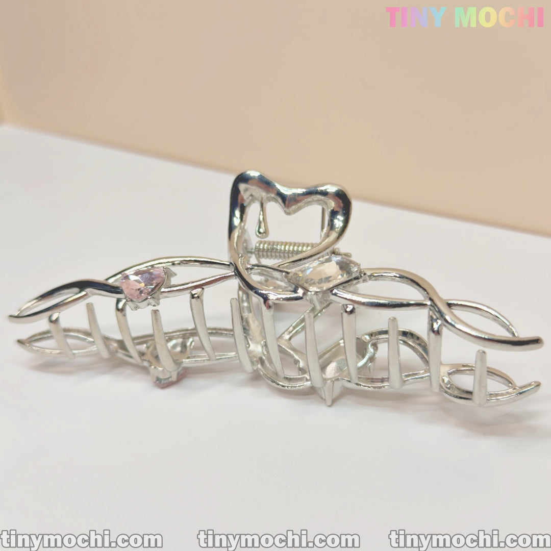 The Tiny Mochi Silver Crystal Heart Hair Claw is an elegant metal hair clip for women, featuring heart details and a cage-like design for a strong grip. Displayed on a light surface, the branded logo is visible in the top right corner.