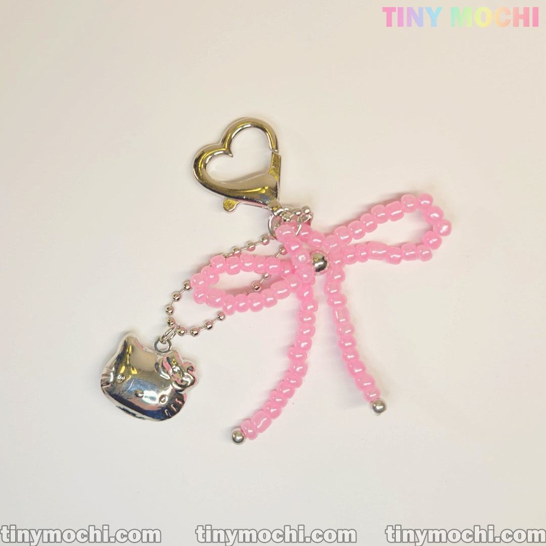 The Pink Beaded Bow & Kitty Charm—Cute Keychain Bag Charm for Coquette Aesthetic by Tiny Mochi features a gold heart-shaped clasp, pink beaded bow, and silver kitty charm. The image shows the Tiny Mochi logo in pastel at the top right corner.