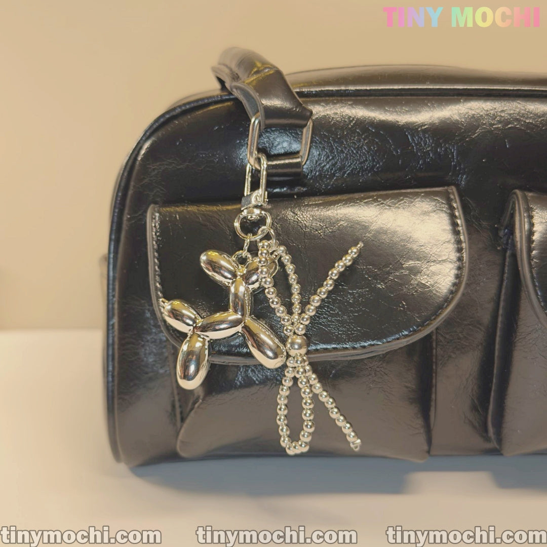 A shiny black handbag with two front pockets is styled with Tiny Mochi’s Silver Balloon Dog Keychain with Bead Bow, adding a trendy touch. The words TINYMochi and tinymochi.com are visible in the image.