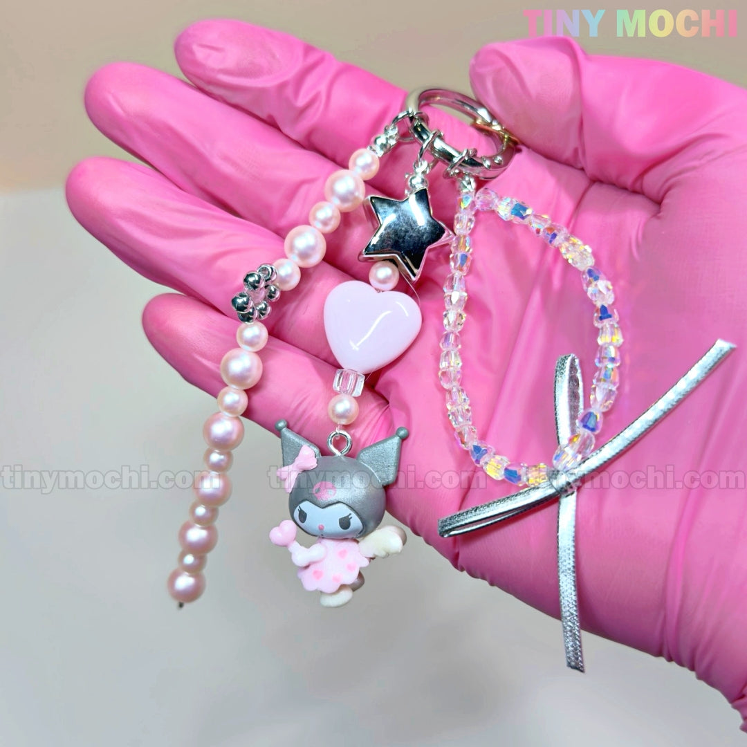 A hand in a pink glove holds the Tiny Mochi Kawaii Angel Charm Keychain Kit, a Sanrio-inspired DIY set with pearls, ribbon, star, pink heart, My Melody charm, and beaded bracelet—perfect for making cute phone or bag accessories.
