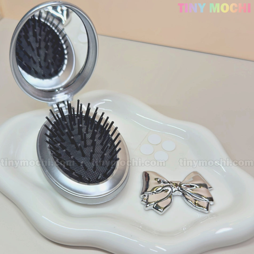 A compact Oval Silver Folding Travel Mirror Comb by Tiny Mochi sits on a white decorative tray beside a shiny silver bow, with the brand name in pastel colors at the top right.