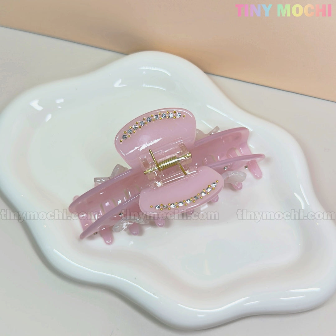 The Pink Bow Claw Clip by Tiny Mochi, made from eco-friendly acetate and adorned with rhinestones and 3D bows, sits on a wavy white ceramic dish. A soft beige background features TINY MOCHI in colorful letters at the top right.