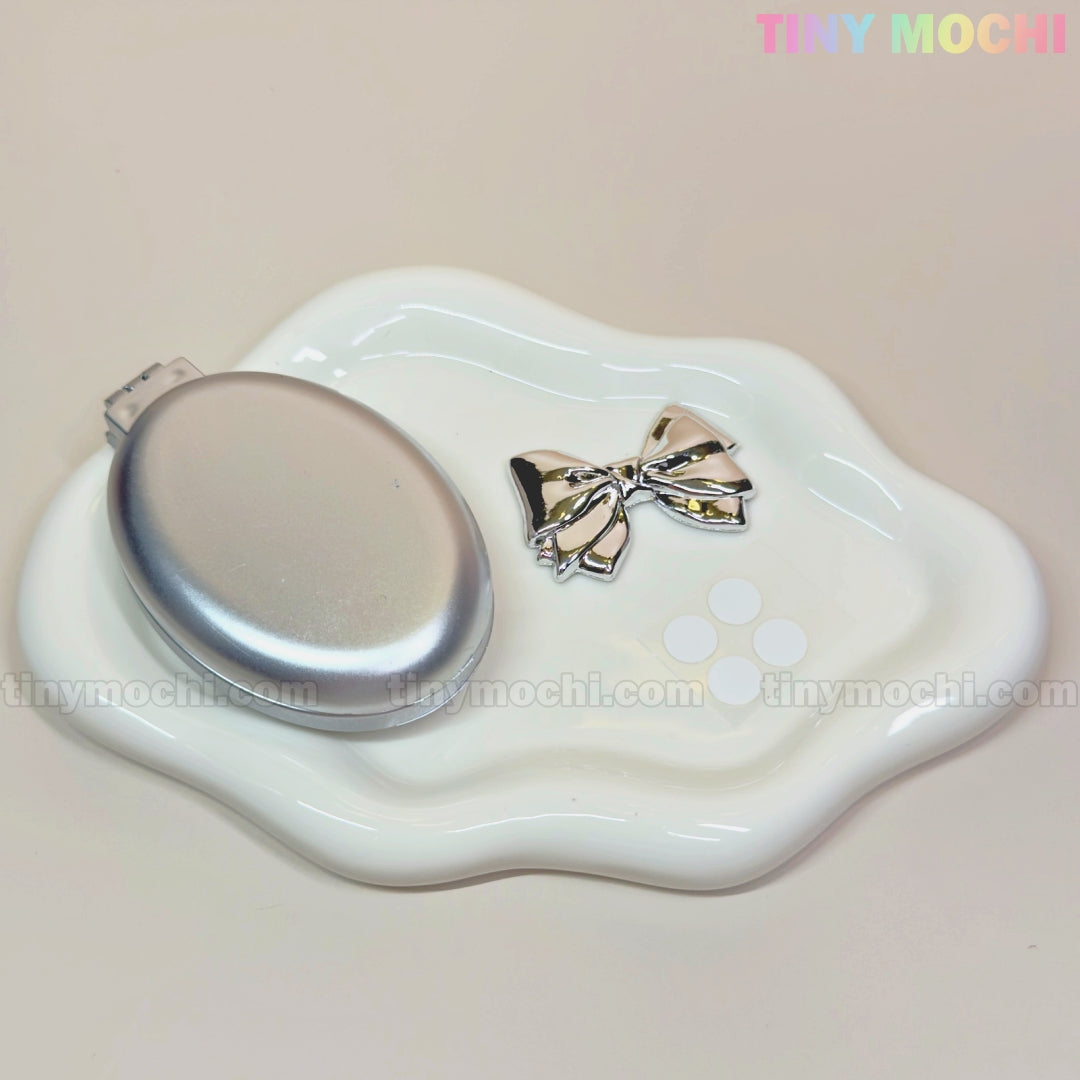 A Tiny Mochi Oval Silver Folding Travel Mirror Comb and a metallic bow hair clip sit on a wavy white ceramic dish. The pastel Tiny Mochi logo shines at the top right, spotlighting this compact 2-in-1 travel essential.
