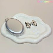 A Tiny Mochi Oval Silver Folding Travel Mirror Comb and a metallic bow hair clip sit on a wavy white ceramic dish. The pastel Tiny Mochi logo shines at the top right, spotlighting this compact 2-in-1 travel essential.