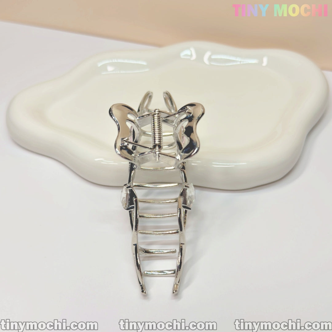 The Silver Crystal Heart Hair Claw by Tiny Mochi, an elegant metal butterfly hair clip for women, shines on a wavy white ceramic dish. A soft beige background and Tiny Mochi in pastel colors highlight the brand at the top right.