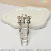 The Silver Crystal Heart Hair Claw by Tiny Mochi, an elegant metal butterfly hair clip for women, shines on a wavy white ceramic dish. A soft beige background and Tiny Mochi in pastel colors highlight the brand at the top right.