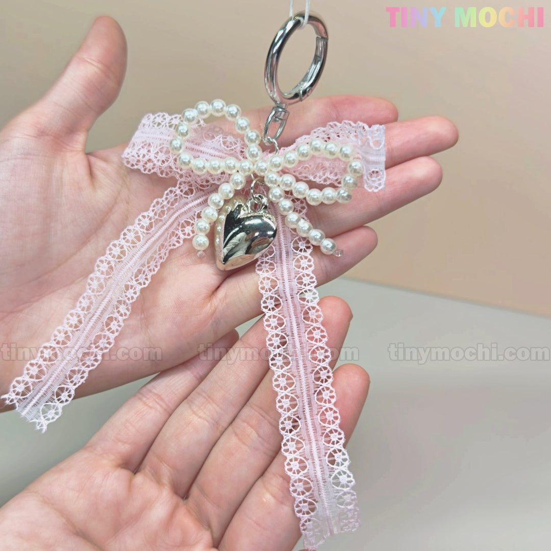 A hand holds the Kawaii Lace Bow Keychain with Pearl Beads & Silver Heart Charm by Tiny Mochiâan adorable bag charm, phone strap, or car mirror accessory. Another hand appears below, with the Tiny Mochi logo in the top right corner.