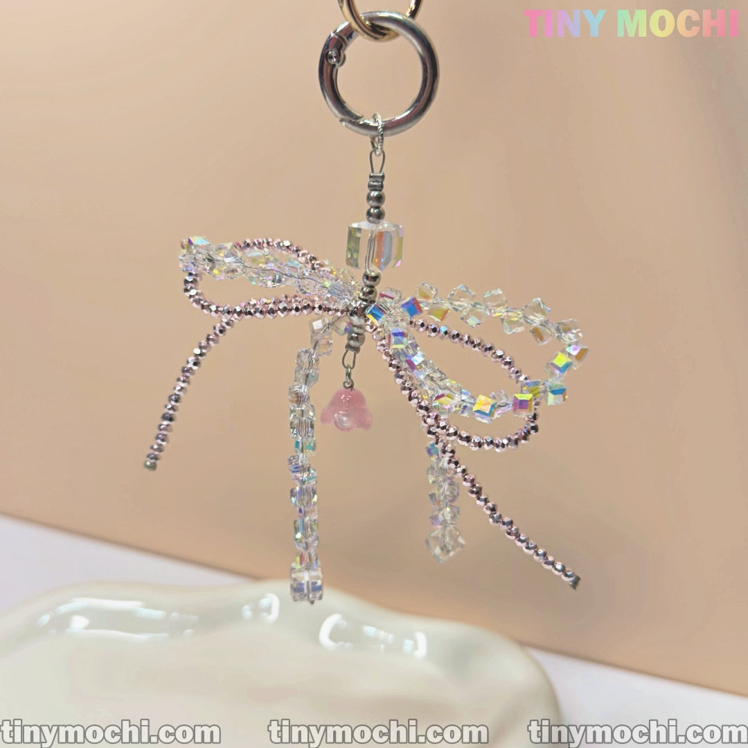 The Tiny Mochi Iridescent Crystal Bow Charm is a delicate keychain with sparkling crystals and a pink bead, perfect for fairycore fans to accessorize bags or phones. Branding appears in the top right corner.