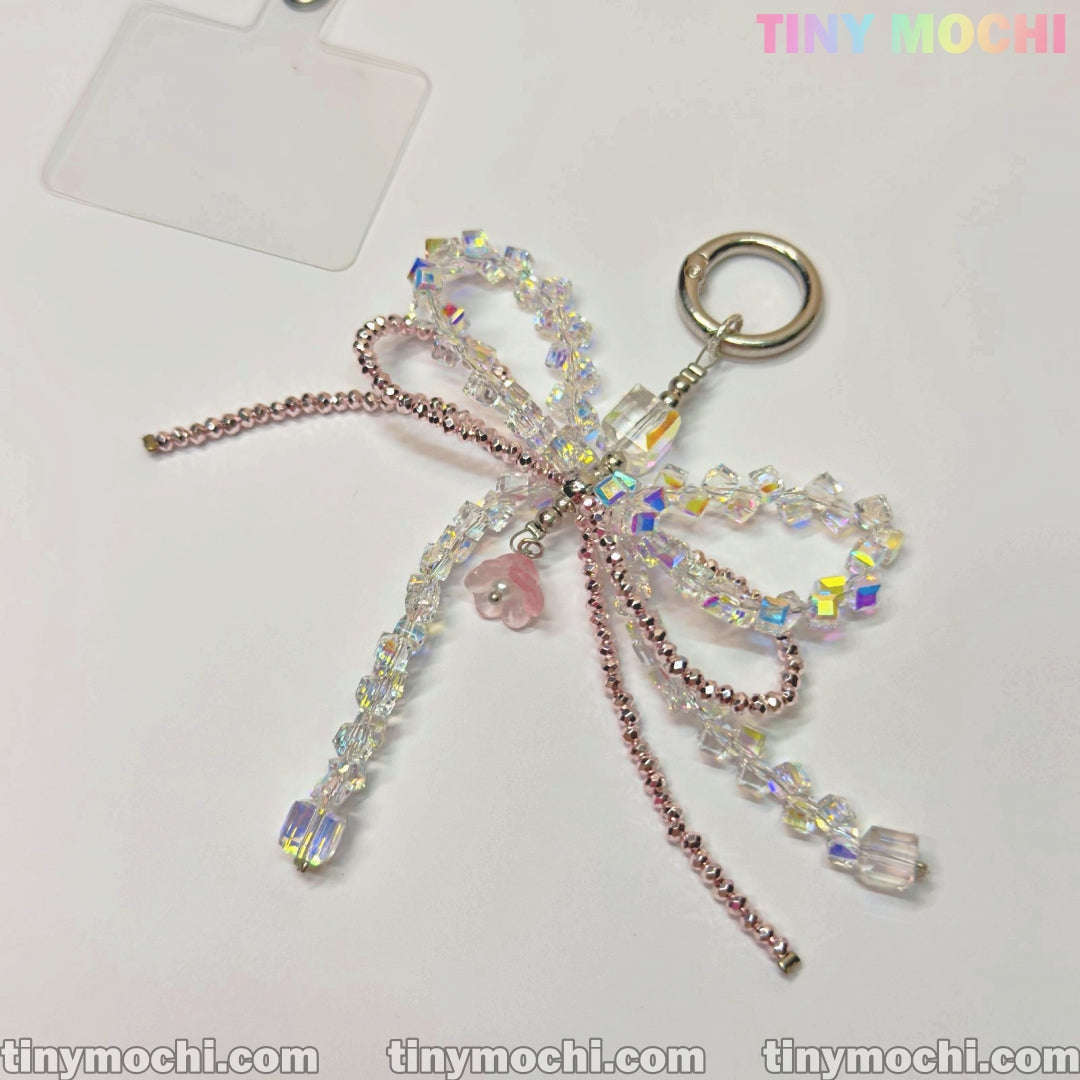 The Iridescent Crystal Bow Charm by Tiny Mochi features clear and pink crystals with a pink bear center, embodying Y2K style. Shown on a white background with the Tiny Mochi logo in the top right corner. Perfect for bags and phones.