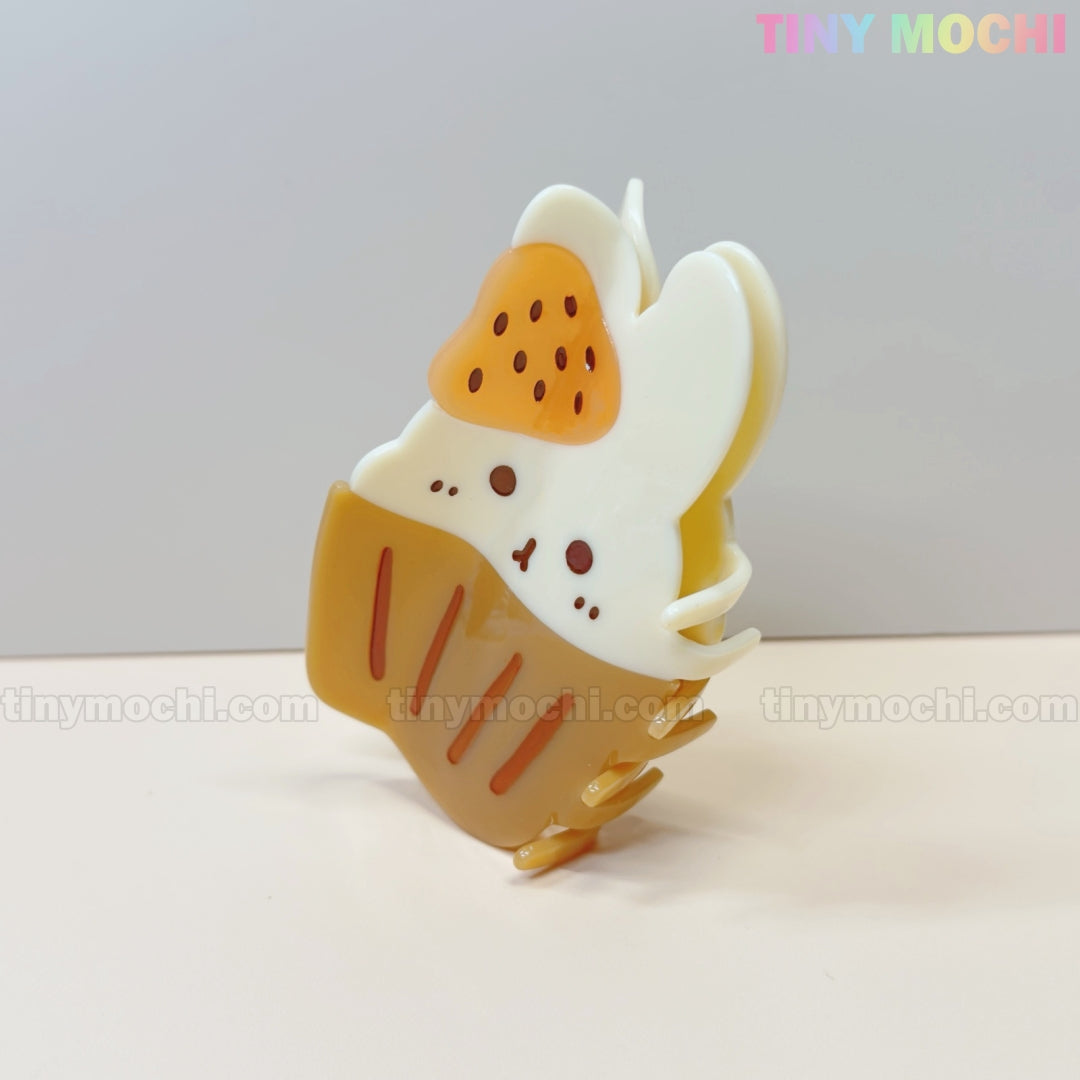 The Kawaii Cupcake Bunny Hair Claw Clip by Tiny Mochi features a bunny in a cupcake with white icing, an orange strawberry on its head, and a brown wrapper—an adorable acrylic grip accessory for women & girls.