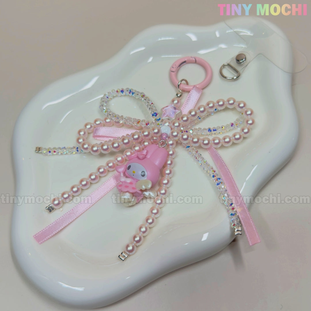 The Tiny Mochi Kawaii Bow Charm Keychain features pink pearls, a ribbon, and a pastel My Melody charm. This cute Coquette Angel bag or phone accessory comes with a clip and is displayed on a white wavy tray.