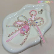 The Tiny Mochi Kawaii Bow Charm Keychain features pink pearls, a ribbon, and a pastel My Melody charm. This cute Coquette Angel bag or phone accessory comes with a clip and is displayed on a white wavy tray.