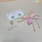 The Tiny Mochi Kawaii Bow Charm Keychain with pearl, ribbon, and coquette angel charm sits beside a transparent clip and business card with QR codes. The background shows the TINY MOCHI logo—ideal as a cute Sanrio-inspired accessory!.
