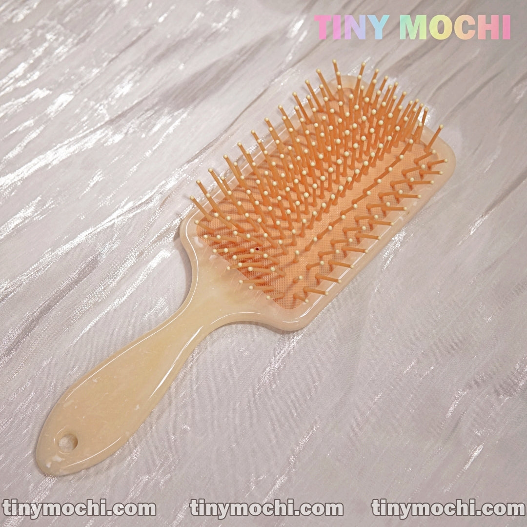 A Marble Pattern Detangling Brush by Tiny Mochi, styled in Nordic fashion for dry and wet hair, with beige bristles, rests on a shiny textured surface. Tiny Mochi appears in pastel colors at the top right corner.