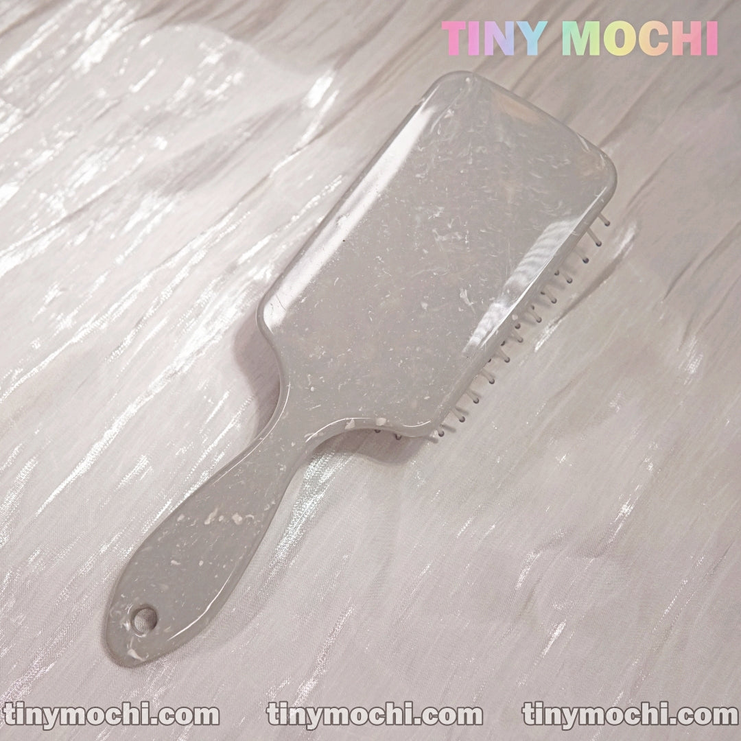A pale, rectangular Marble Pattern Detangling Brush by Tiny Mochi rests on a shiny marble-patterned surface. Tiny Mochi appears in pastel in the top right, with tinymochi.com repeated below. Suitable for dry and wet hair.