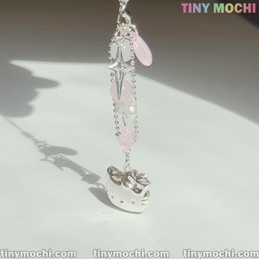 A Pink Kitty Phone Charm from Tiny Mochi, featuring kawaii Y2K coquette style with pink and clear beads, hangs from a chain on a white surface. TINYMochi.com and TINY MOCHI are visible in the image.