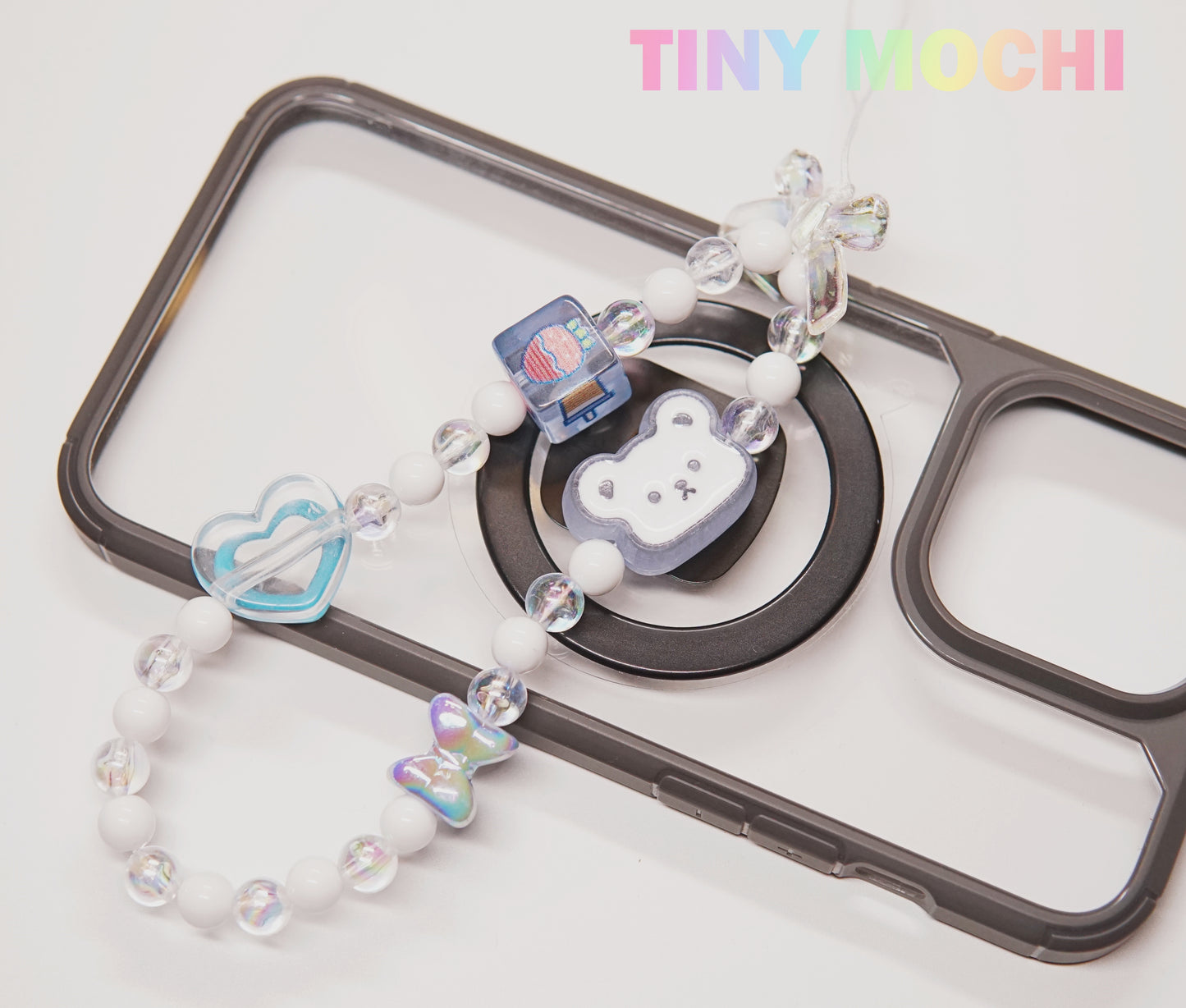 The Y2K Aesthetic Phone Charms Set by Tiny Mochi, featuring bear, butterfly, and heart charms on colorful lanyards, hangs over a black and clear case. “Tiny Mochi” appears in pastel at the top right, adding nostalgic vibes.