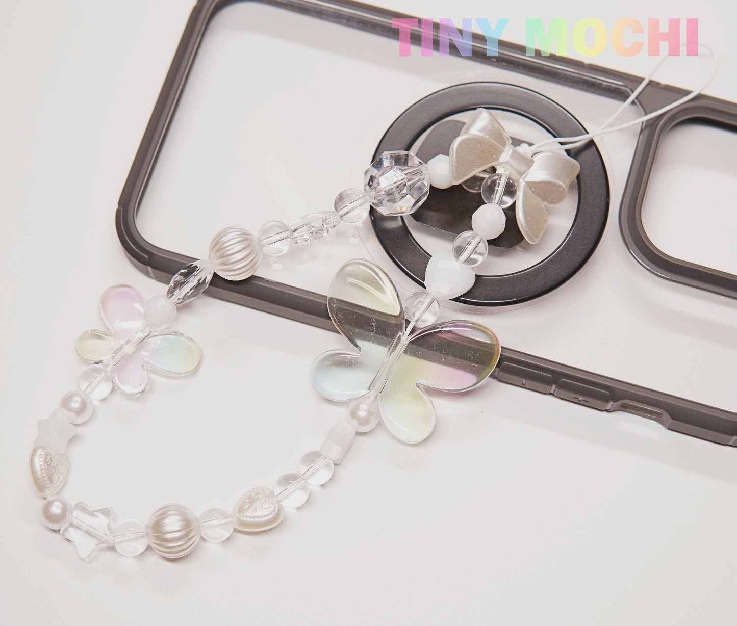 The Y2K Aesthetic Phone Charms Set from Tiny Mochi features bear, butterfly, and heart charms on colorful lanyards, adding playful vibes to your black phone case. The Tiny Mochi brand name appears in colorful letters in the top right corner.