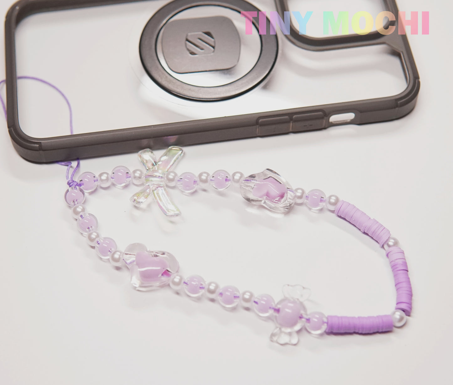 The Y2K Inspired Beaded Phone Strap by Tiny Mochi, featuring butterflies and hearts, is shown beside a black phone case on a white background. TINY MOCHI appears in colorful letters at the top right. Ideal for phones, AirPods, and more!.