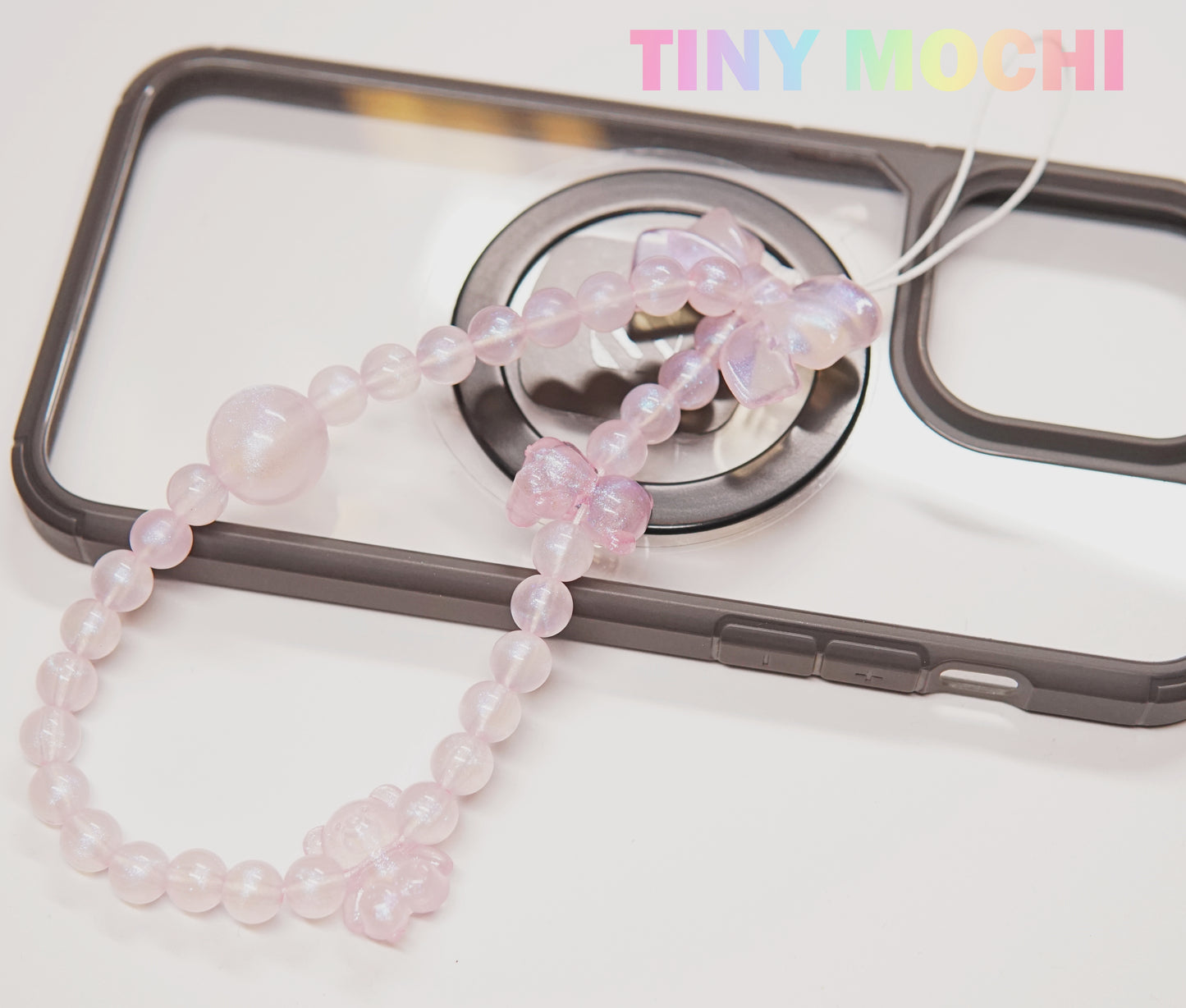The Tiny Mochi Y2K Inspired Beaded Phone Strap Collection features pink butterfly and round beads, adding a trendy touch to black or clear phone cases. TINY MOCHI appears in pastel rainbow letters in the top right corner.