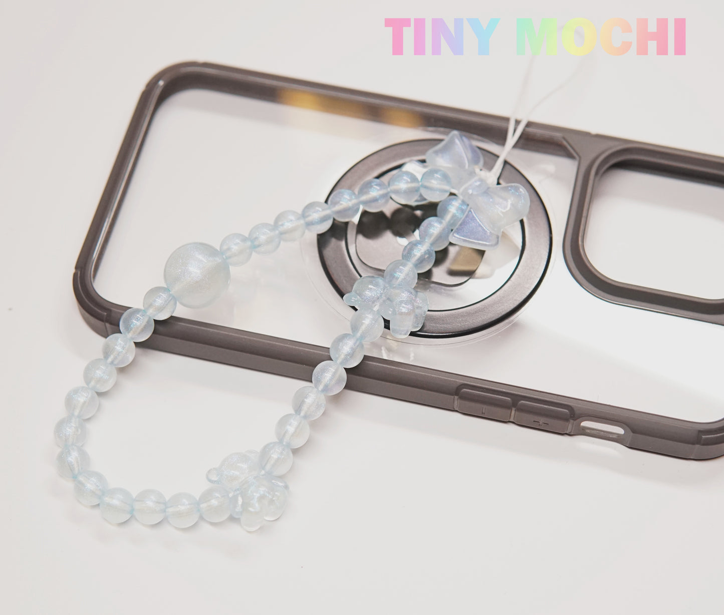 A clear phone case with a ring grip is decorated with a translucent blue strap from the Tiny Mochi Y2K Inspired Beaded Phone Strap Collection, adding fun charms to your daily essentials. The Tiny Mochi name appears in pastel colors for a trendy touch.