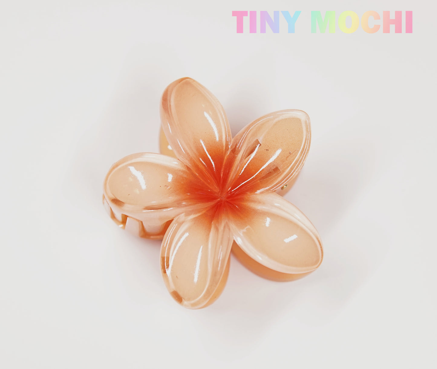 The Plumeria Flower Hair Claw by Tiny Mochi, a stylish floral hair accessory in peach, is displayed on a white background with TINY MOCHI in pastel rainbow text at the top right corner.