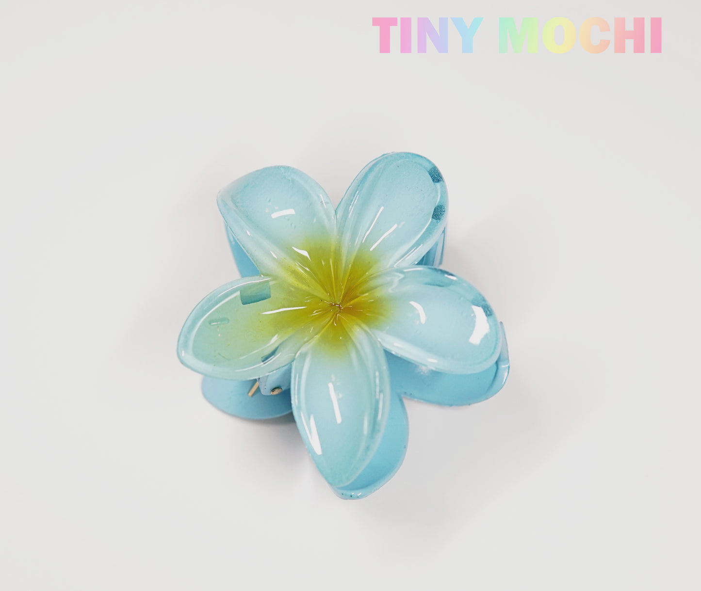 The Tiny Mochi Plumeria Flower Hair Claw is a stylish floral hair accessory in shiny blue with a yellow center, shown on a white background. TINY MOCHI appears in pastel colors, highlighting this cute hair clip for women and girls.