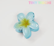 The Tiny Mochi Plumeria Flower Hair Claw is a stylish floral hair accessory in shiny blue with a yellow center, shown on a white background. TINY MOCHI appears in pastel colors, highlighting this cute hair clip for women and girls.
