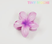 The Plumeria Flower Hair Claw from Tiny Mochi, a glossy light purple floral hair clip with a darker purple center, is displayed on a white background. The TINY MOCHI logo appears in pastel rainbow colors in the top right corner.