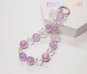 The Tiny Mochi Adorable Faux Pearl Bag Charm Keyring features iridescent purple and clear beads, a clear plastic bow, and pearl accents. Styled as a whimsical phone chain, its shown on a white surface with “TINY MOCHI” in pastel at the top right.