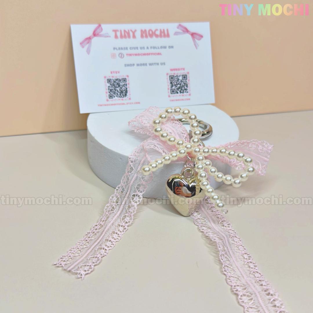 The Tiny Mochi Kawaii Lace Bow Keychain with pearl beads and a silver heart charm sits on a round white box, with a Tiny Mochi business card behind it against a beige backdrop.