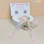 The Tiny Mochi Kawaii Lace Bow Keychain with pearl beads and a silver heart charm sits on a round white box, with a Tiny Mochi business card behind it against a beige backdrop.